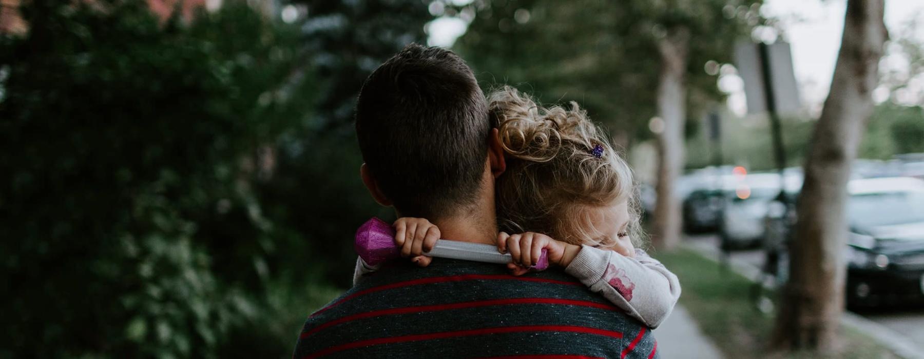 tired little girl rests her head on her father's shoulder as he carries her down the city sidewalk